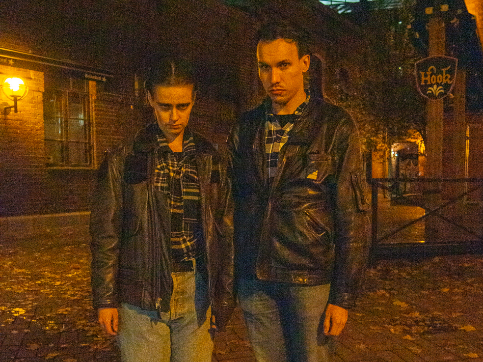 Kaksi miesdragiin pukeutunutta seisoo vierekkäin ja katsoo kameraan valaistulla kujalla